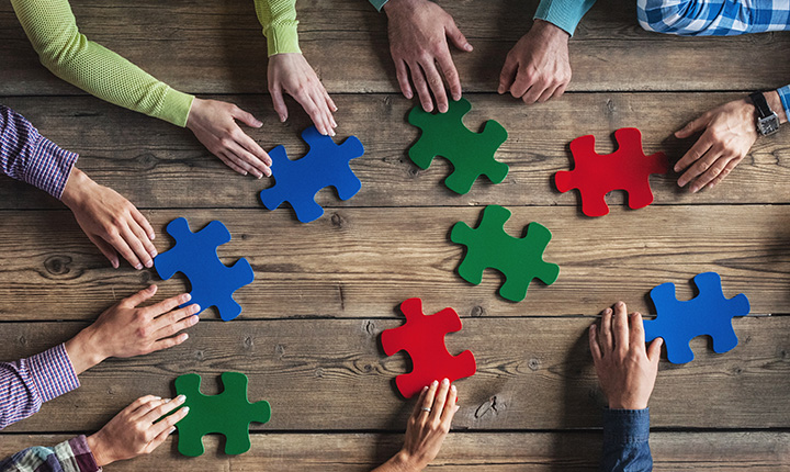 Hands around a wooden table holding red, blue and green puzzle pieces