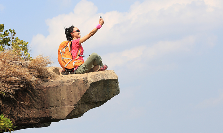 Hiker sitting on a rock cliff taking a photo with a phone
