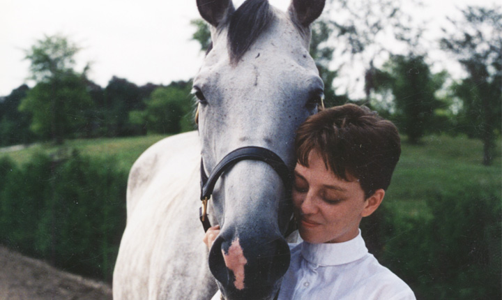 A woman wearing a white collared shirt with her face beside a grey horse.