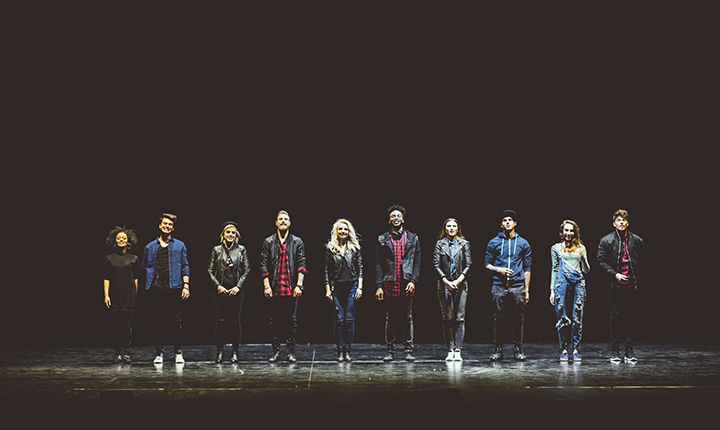 A diverse group of actors standing on a black stage with a spotlight shining on them 